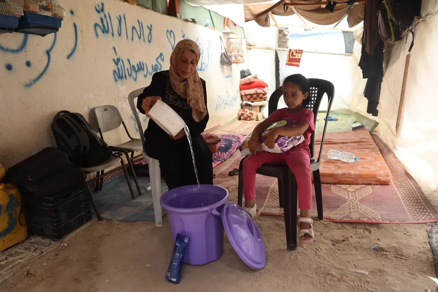 A woman pours water into a large purple bucket beside a seated child inside a makeshift shelter.
