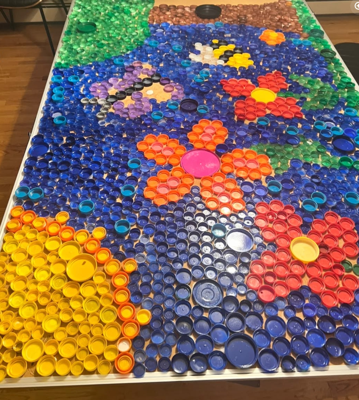 Completed bottle-cap mural showing flowers, a butterfly, and greenery made from thousands of colored caps