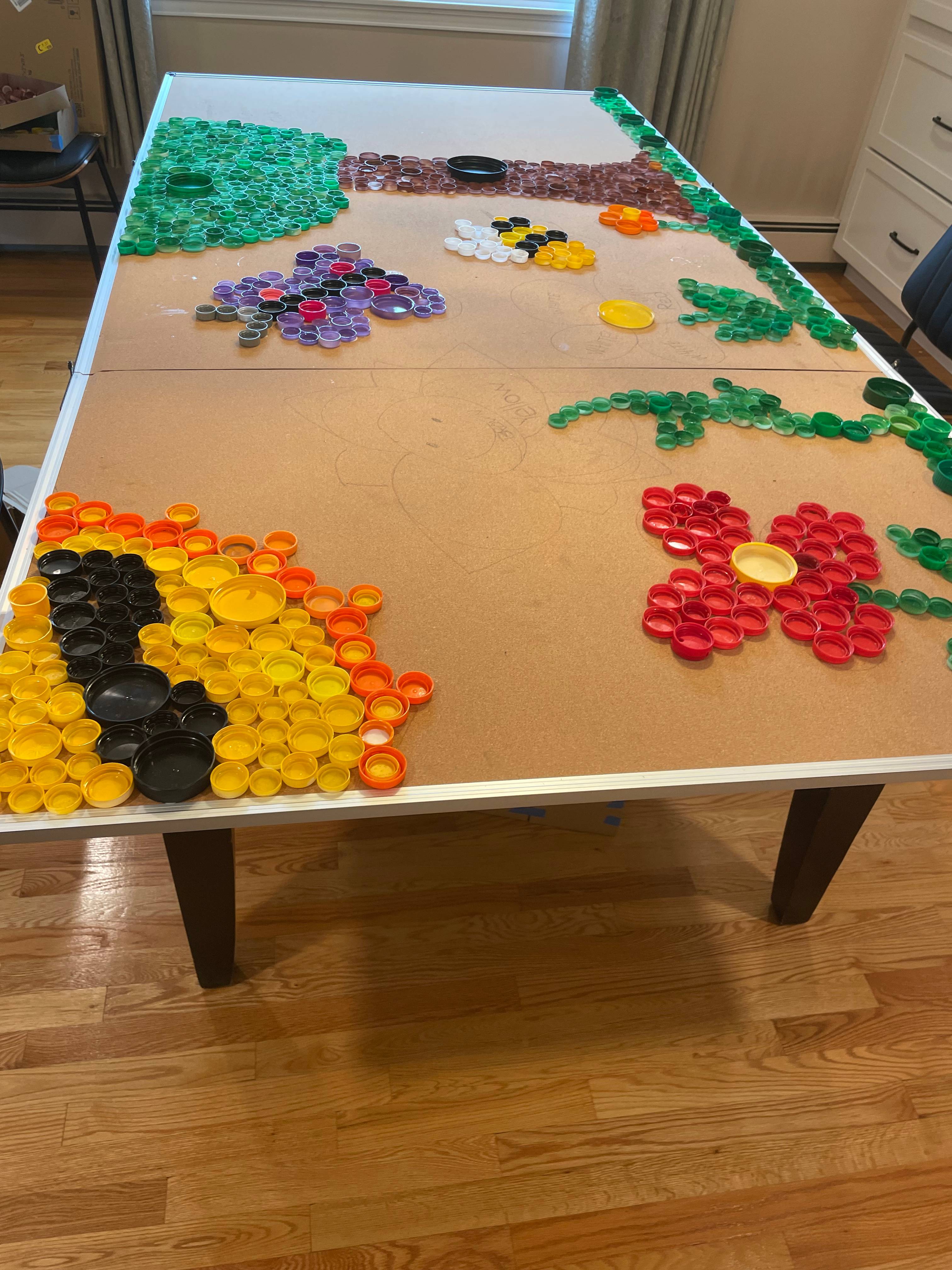 Bottle-cap mural in progress showing a tree, flowers, and vines made from colored caps